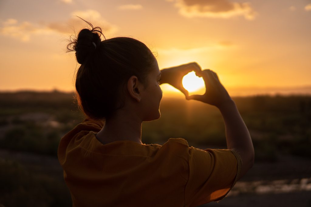 young lady makes heart with hands around sun glare