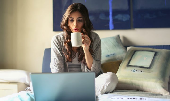 woman-in-grey-jacket-sits-on-bed-uses-grey-laptop